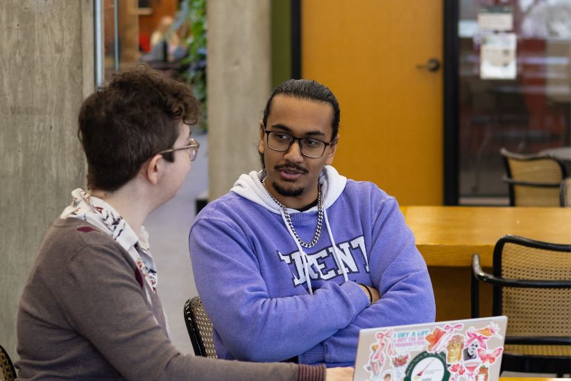 A student and a Librarian sit at a table having a conversation, with one wearing a Trent University hoodie and a laptop covered in colorful stickers in front of them.