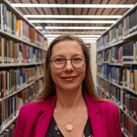 A woman with long straight hair and glasses, wearing a bright pink blazer over a floral top, smiles in a library.