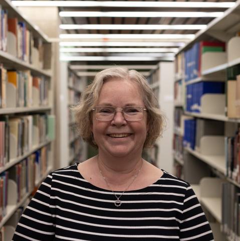 A woman with glasses and short hair smiles in a library, wearing a black and white striped shirt.