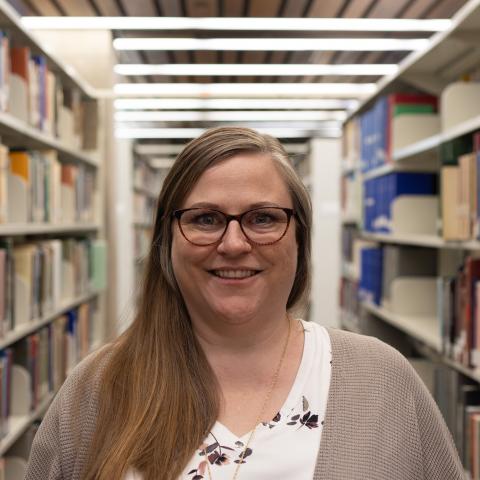 A woman with long brown hair and glasses smiles in a library, wearing a white floral top under a light cardigan.
