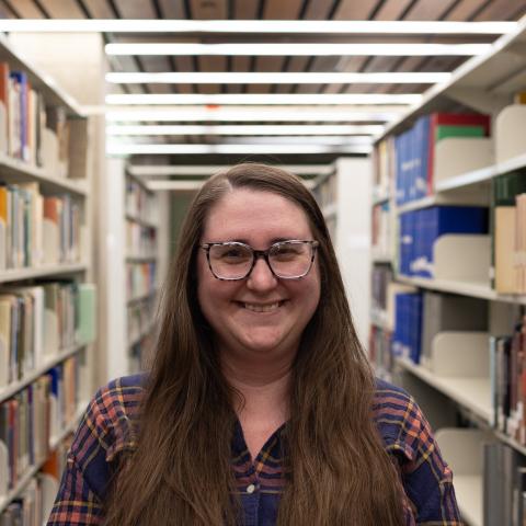 A woman with long brown hair and glasses smiles in a library, wearing a plaid shirt.
