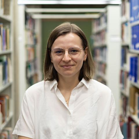 A woman with shoulder-length brown hair, glasses, and a white shirt is standing in a library aisle, smiling slightly at the camera.